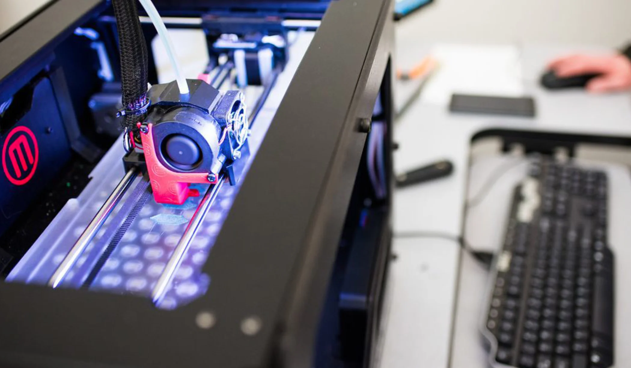 A 3D printer in operation, with its print head moving along metal rails as it builds an object. The machine’s interior is lit with a cool blue light, and a person works at a nearby computer workstation with a keyboard and mouse visible in the foreground.
